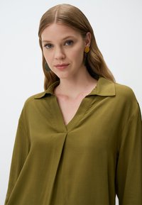 Blusa verde oliva con scollatura a V e maniche lunghe, realizzata in tessuto morbido caratterizzato da un leggero drappeggio, che mette in evidenza una vestibilità comoda.