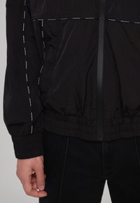 Schwarze Windjacke mit elastischen Bündchen und Saum, ausgestattet mit einem Reißverschluss und reflektierenden Details entlang der Nähte.