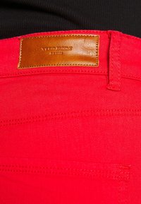 Jeans de mezclilla rojos con una etiqueta de cuero marrón. La tela tiene una textura suave y costuras visibles a lo largo de las costuras.