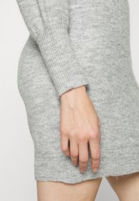 Szara dzianinowa sukienka z prążkowanymi mankietami, o lekko teksturowanej powierzchni i długości kończącej się powyżej kolan. Ręka umieszczona na boku.