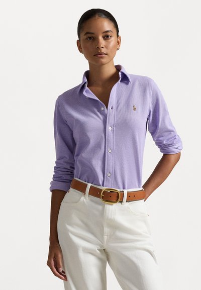 Cămașă lungă cu mânecă, de culoare violet deschis, cu guler tip butonat, prezentând un mic logo brodat, asortată cu pantaloni de culoare crem și o curea maro.