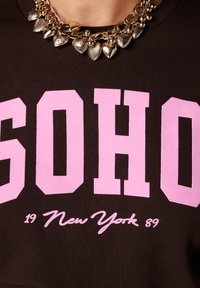 Bruine sweatshirt met gedurfde roze blokletters die "SOHO" en "New York 89" spellen, aangevuld met een opvallende gouden halsketting met hartvormige hanger.