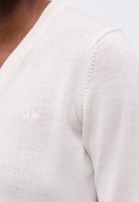 Κοντινό πλάνο ενός ατόμου που φοράει λευκό πουλόβερ πλεκτό με V-λαιμόκοψη και μικρό κεντημένο λογότυπο στο στήθος.