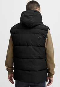 Gilet imbottito nero con cappuccio, caratterizzato da cuciture orizzontali e un piccolo logo sul retro. Indossato sopra una camicia a maniche lunghe marrone.
