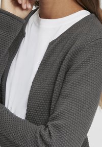 Gray knit cardigan with a textured surface and open front, layered over a white t-shirt, featuring a relaxed fit and long sleeves.