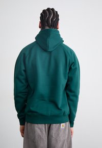 Dark green hoodie with a kangaroo pocket, ribbed cuffs, and hem. Smooth texture, featuring a large hood. Worn with gray corduroy pants.
