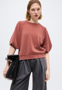 Γυναίκα ντυμένη με ροζ σκόπιμο κοντομάνικο τοπ, γκρι πλισέ παντελόνια, κρατώντας μια μεγάλη μαύρη τσάντα φάκελο μπροστά σε απλό φόντο.