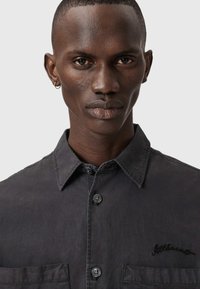 Primo piano di un uomo con pelle scura che indossa una camicia a bottoni grigio scuro con due tasche frontali e un piccolo logo ricamato.