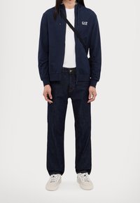 Marinblå zip-up hoodie med vit logotyp, kombinerad med lösa mörka denimjeans och vita sneakers. Mjukt tyg med en avslappnad design.