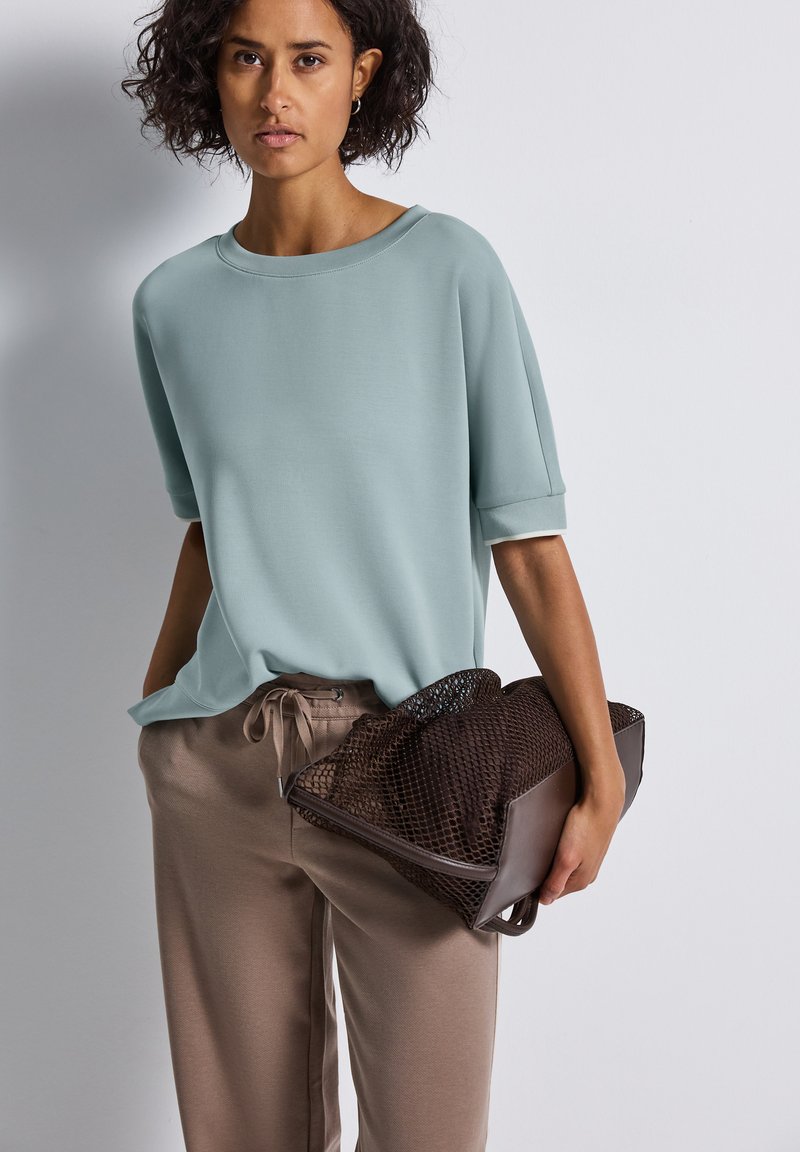 Haut bleu clair à manches courtes associé à un pantalon beige avec cordon de serrage, tenant un sac en maille marron texturé, présentant une finition en tissu lisse.