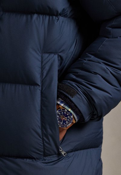 Tengerészkék steppelt dzseki mellzsebbel, tépőzáras rögzítéssel. Alatta kockás ing látható, kék számlapos faórával.