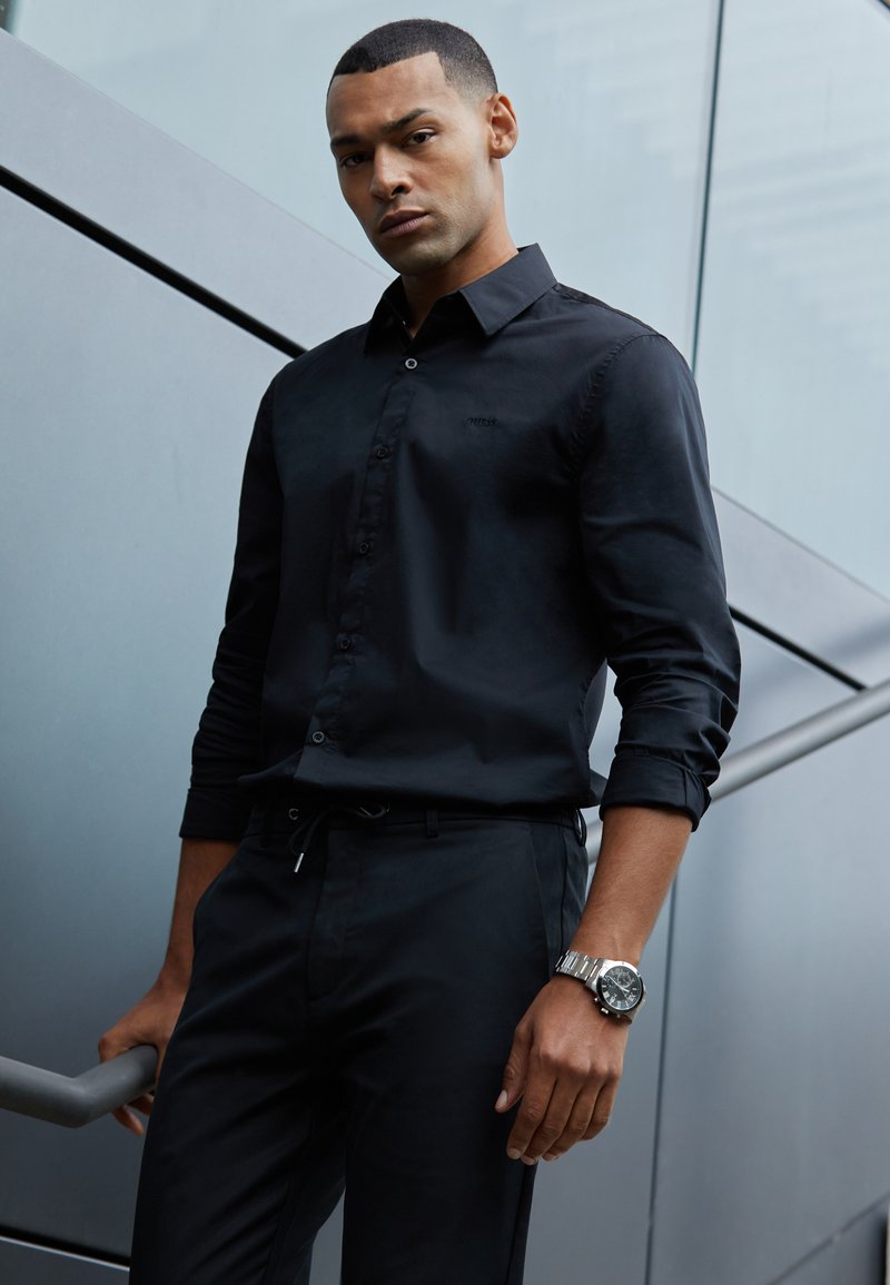 Chemise noire à boutons avec une texture lisse, associée à un pantalon noir. Montre en argent notable au poignet. Le modèle se tient devant un fond gris.