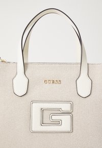 Sac fourre-tout beige texturé avec des accents en cuir blanc, logo "GUESS" doré, et un grand logo en forme de G blanc à l'avant. Double poignées supérieures.