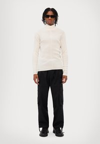 Gai bēša ribota turtleneck džemperis, melnas cargo bikses ar kabatām un melnas kurpes. Modelis valkā tumšus saulesbrilles un stāv pretī vienkāršai fona.