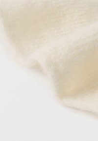 Tkanina v jemnej krémovej farbe s chlpatou textúrou a jemným vzorom, jemne zavesená, aby ukázala svoju tvárnosť a ľahkú váhu.