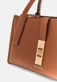 Borsa a mano in pelle marrone con design strutturato, un accento dorato con logo del marchio e una tracolla removibile per diverse opzioni di trasporto.