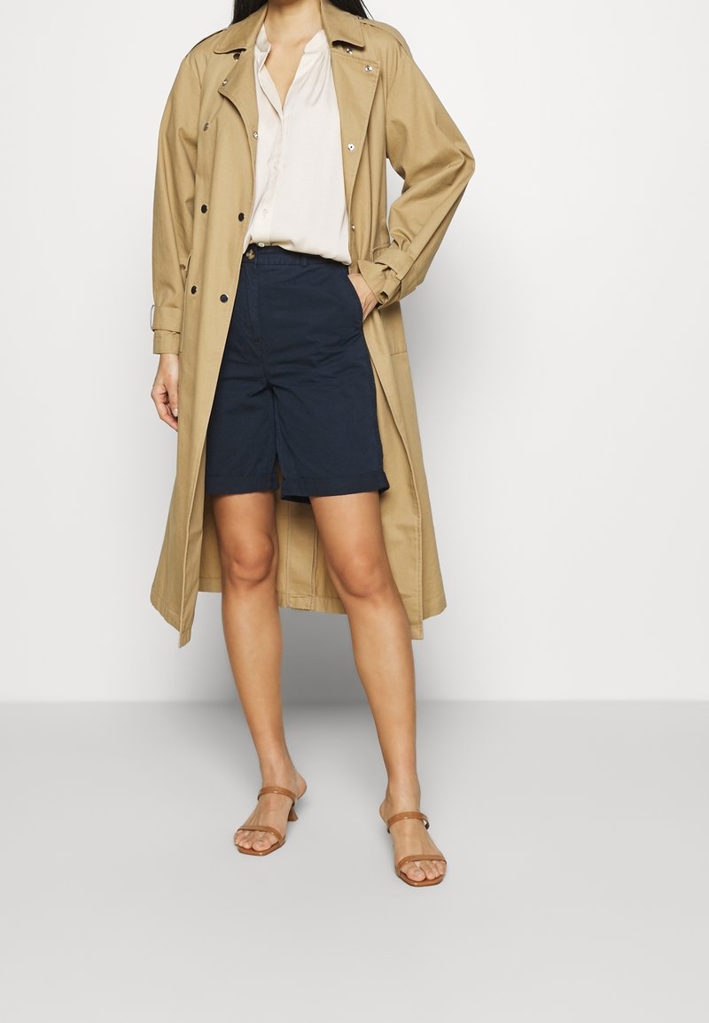 Femme portant un trench-coat beige, un chemisier blanc, un short marine à hauteur des genoux, et des sandales à talons marron clair, debout devant un fond clair uni.