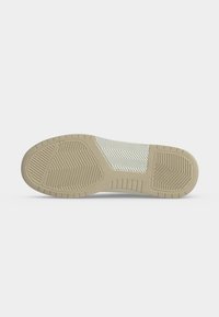 Semelle d'une basket beige et blanche avec des motifs de grip texturés au talon, à la semelle intermédiaire et à l'avant du pied sur un fond gris clair uni.