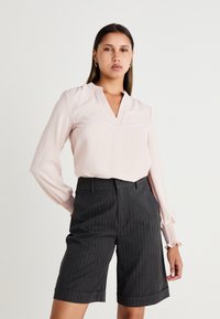 Helle rosa Bluse mit langen Ärmeln, V-Ausschnitt und Puffärmeln, kombiniert mit dunkelgrauen taillierten Shorts mit Nadelstreifen und umgeschlagenem Saum.