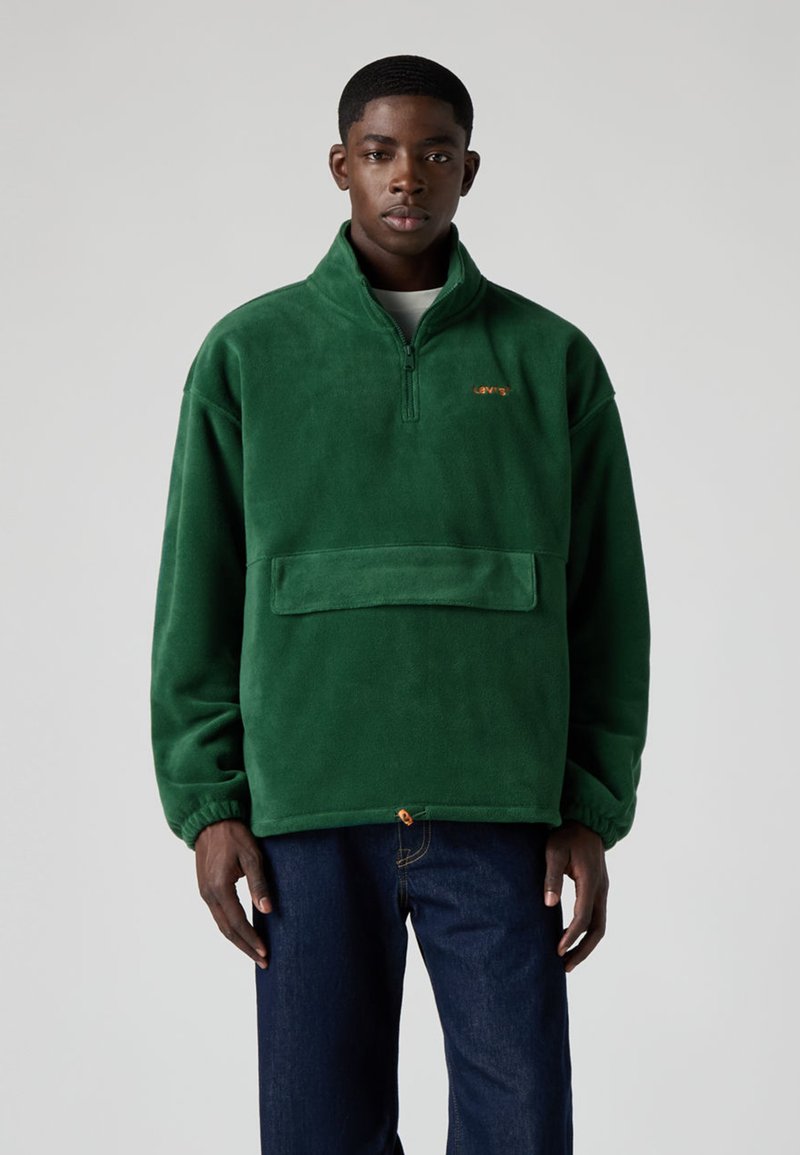 Grön fleecetröja med halvlång dragkedja vid kragen, framficka, elastiska ärmslut och kontrasterande orange logotyp på bröstet. Bärs med mörka jeans.