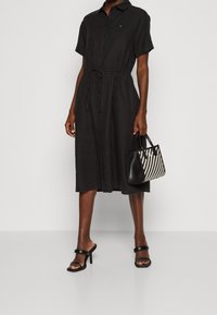 Černé košilové šaty s krátkými rukávy, se stažením v pase a střední délkou. Doplňují je malá černobílá kostkovaná kabelka a sandály na jehlovém podpatku.