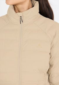 Beige, vadderad jacka med hög krage, framdragkedja och diskret logotypdetalj. Materialet verkar slätt med en lätt design.