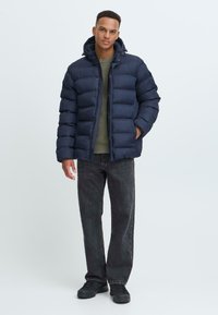 Marineblå dunstjakke med glidelås og knappelukking, med hette og kviltet mønster. Kombinert med mørke jeans og svarte sko.