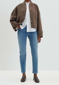 Ruskea lyhyt takki, jossa edessä napit, valkoinen paita, vaaleansiniset slim-fit-farkut ja tummanruskeat slip-on-kengät, neutraalia taustaa vasten.