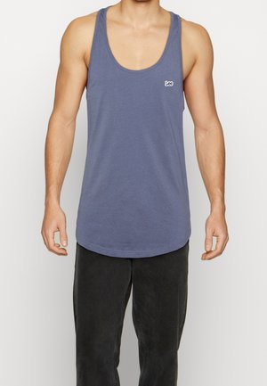 Homme portant un débardeur bleu sans manches avec le logo « Lee » sur la poitrine et un pantalon noir, debout devant un fond uni.