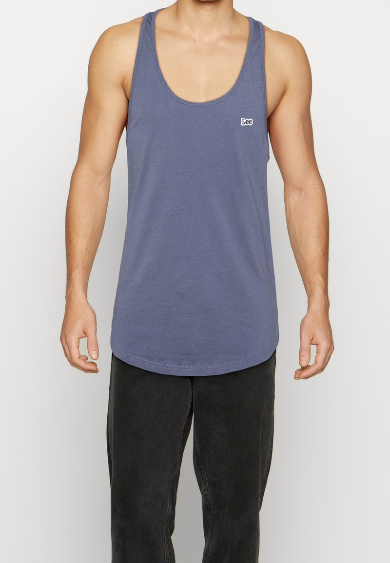 Homme portant un débardeur bleu sans manches avec le logo « Lee » sur la poitrine et un pantalon noir, debout devant un fond uni.