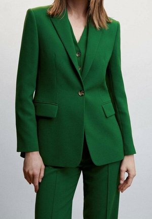 Femme portant un tailleur vert foncé sur mesure avec une veste boutonnée une fois et un pantalon assorti, debout devant un fond uni.