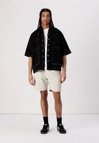 Svart oversized skjorte med hvite sømmer over en hvit t-skjorte; kombinert med beige shorts og svarte sneakers med hvite detaljer.