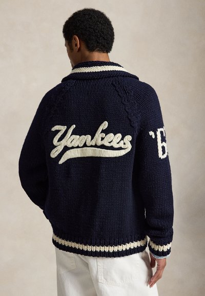 Homme portant une veste en maille bleu marine avec le texte blanc « Yankees » dans le dos et un col et des poignets rayés, associée à un pantalon blanc.