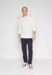 Maglione crema con maniche lunghe, pantaloni blu e sneakers multicolore. Texture morbida e vestibilità comoda, in piedi su uno sfondo neutro.