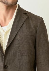 Brauner kariert Blazer mit Reverskragen, einem Einzelknopfverschluss und einer Brusttasche. Getragen über einem hellen, gemusterten Hemd.