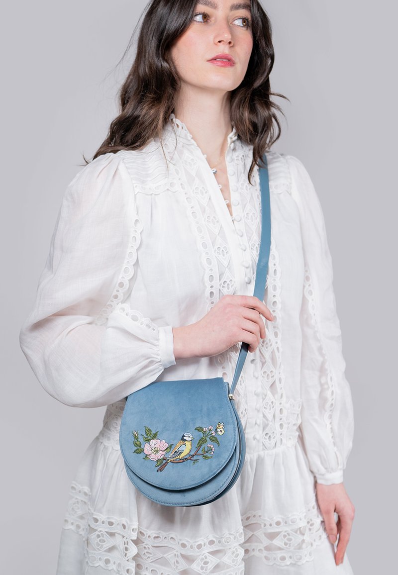 Sac bandoulière rond en suède bleu avec un design brodé d'oiseau et de fleurs. Doté d'une fermeture zippée et d'une bandoulière réglable.