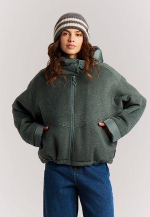 Frau trägt grüne gesteppte Jacke mit Reißverschluss, Hände in den Taschen, gestreifte Strickmütze und blaue Jeans, steht vor einem schlichten Hintergrund.
