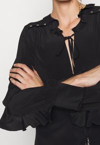 Blouse noire avec col et manchettes volantés, fermeture à nouer à l'avant, boutons brillants sur les épaules, tissu lisse avec un léger éclat.