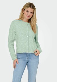 Cardigan texturé vert clair avec un col rond et une fermeture à boutons à l'avant. Assorti à un jean bleu taille haute. Design décontracté et ajusté.