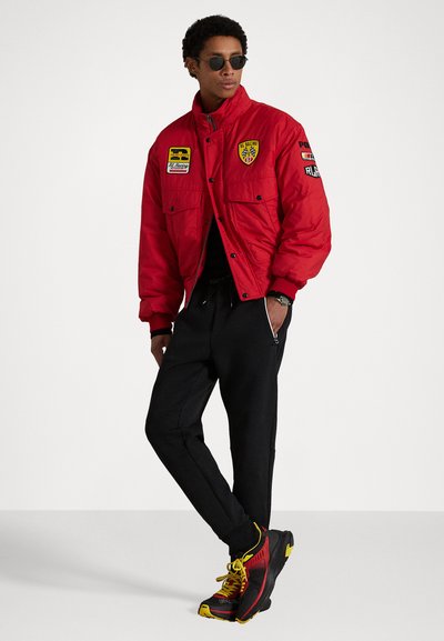 Blouson bomber isolé rouge avec poches avant, orné de patches ; associé à un pantalon de jogging noir et des baskets rouges, noires et jaunes.