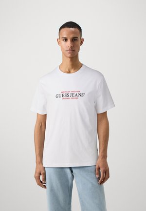 Weißes Baumwoll-T-Shirt mit dem Schriftzug "GUESS JEANS" in Rot und Schwarz. Kurze Ärmel, runder Halsausschnitt und ein lässiger Schnitt.