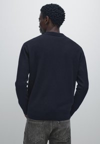 Navy blue knitted sweater with a crew neck, long sleeves, and ribbed cuffs and hem, worn with light gray jeans, viewed from the back.