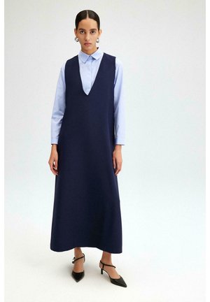 Naine, kes kannab tumesinist varrukateta kleiti helesinise kraega särkidega, mustade teravate kontsadega ja hõbedaste rõngakõrvarõngastega.