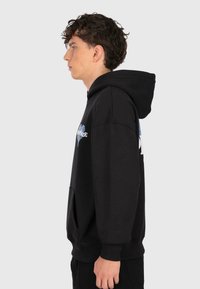 Young man with curly hair wearing a black hoodie with blue and white graphic, standing in profile against a plain light background.