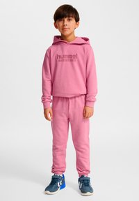 Sudadera con capucha de forro polar rosa con logo frontal y pantalones deportivos a juego. Capucha, puños acanalados, cintura elástica. Zapatillas deportivas azules con detalles.