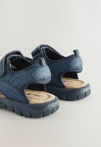 Blå sandaler i perforerat syntetläder, med kardborreband och en texturerad gummisula. Mjuk beigemellanfotsula för komfort.
