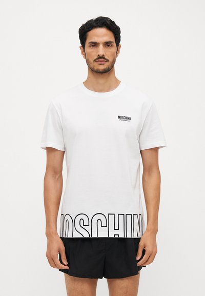 Valkoinen puuvillapaita, jossa on pyöreä kaula-aukko, lyhyet hihat ja iso musta "MOSCHINO"-logo alhaalla. Siisti, minimalistinen muotoilu.