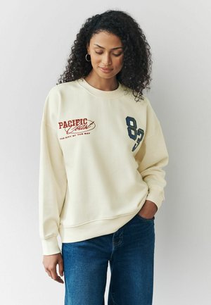Cremfarbener Sweatshirt mit dem Aufdruck "PACIFIC" und der Nummer "83" in dunkelblau. Glatte Textur mit gerippten Bündchen und Saum.