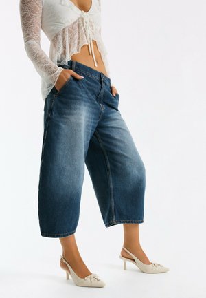 Kvinde iført cropped blå vidde bukser, hvid blonder langærmet cropped top og hvide sandaletter med kitten heel og åben hæl.