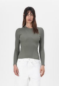 Maglietta a maniche lunghe verde oliva a costine con un design aderente, caratterizzata da dettagli di cucitura sottili e un collo rotondo, abbinata a pantaloni bianchi.